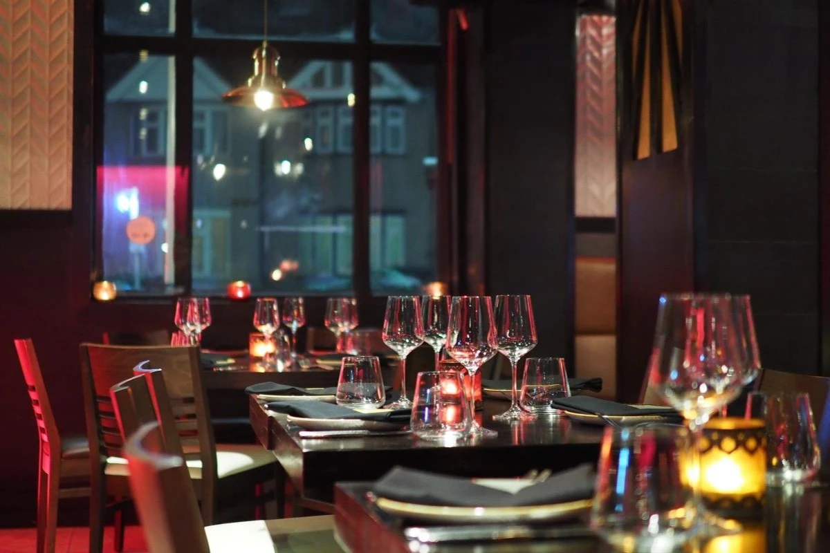 mehrere festliche gedeckte Tische und Sessel in einem gehobenen Restaurant, im Hintergrund ein Fenster, durch das man Häuserfassaden bei Nacht sieht