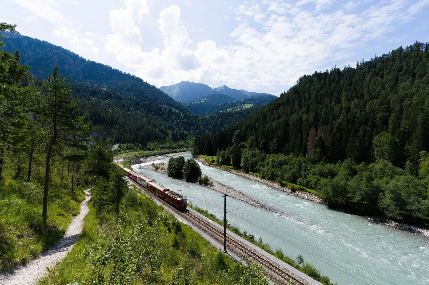 Gebirgstal mit Fluss und daneben liegender Zugsstrecke, zu sehen ist ein durchfahrender Zug